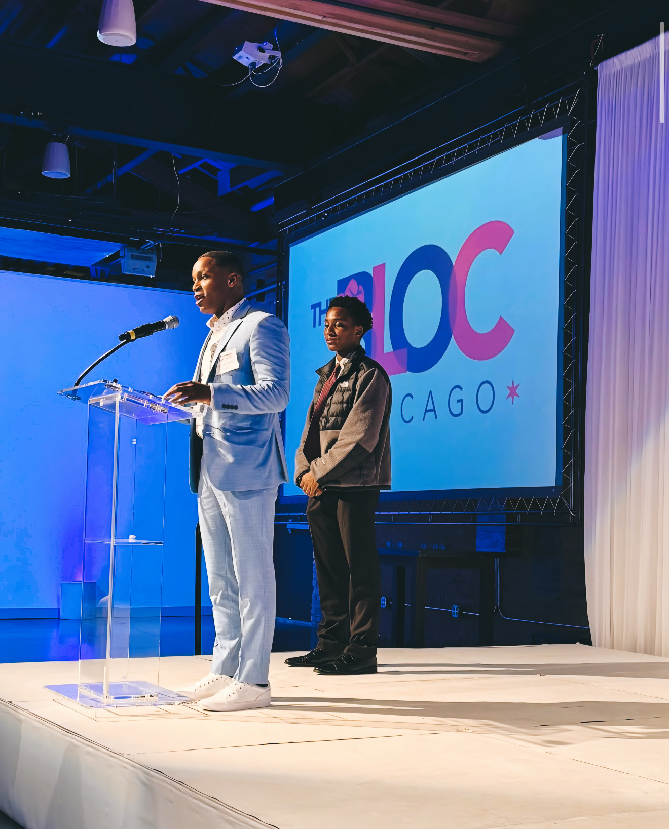 Jamyle Cannon speaking at a podium next to a young Bloc student, with The Bloc Chicago logo projected behind them